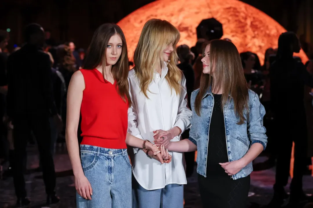 Nicole Kidman shares a warm moment with her daughters, Sunday Rose, and Faith Margaret, while attending the Chanel Womenswear Spring/Summer 2026 show during Paris Fashion Week, on October 6, 2025. The trio stands hand in hand under the soft glow of the event's dramatic lighting.