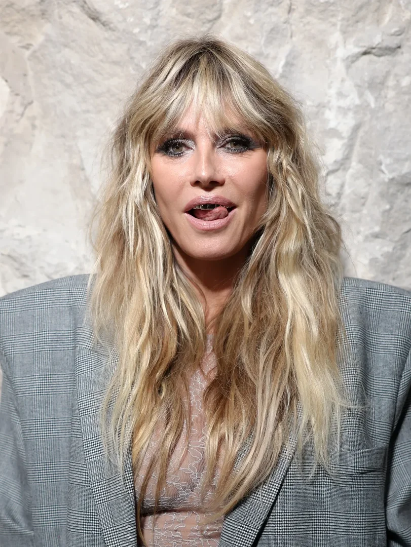Heidi Klum’s daring look goes beyond the sheer dress as the camera zooms in on her face, revealing her silver grillz. She playfully positions her tongue against the upper set, the metallic shine catching the light from the camera flash.