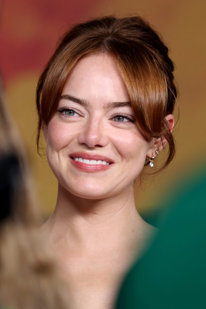 Emma Stone beams with an easy, luminous smile, tilting her head slightly as soft tendrils frame her face — a picture of poise and quiet confidence on the red carpet.