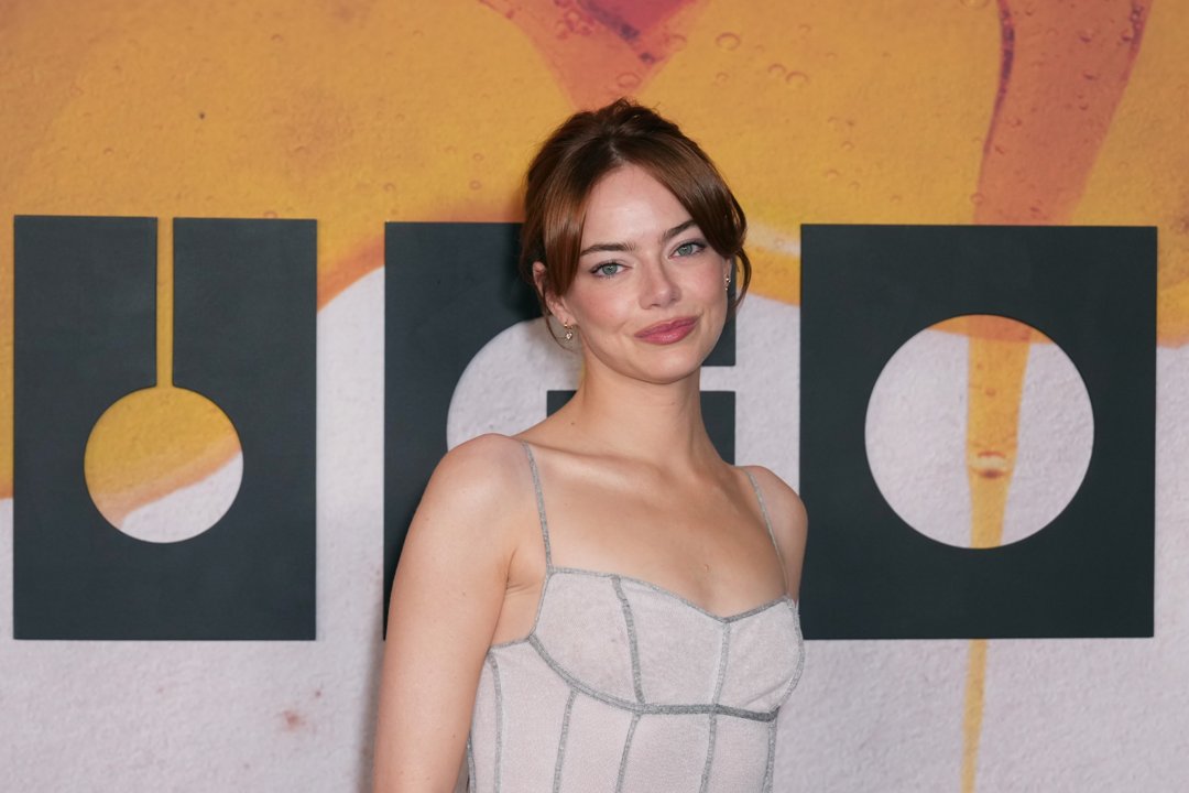Emma Stone poses with confidence, her shoulders angled and lips curved in a knowing half-smile. The soft sheen of her gown catches the light against the bold 