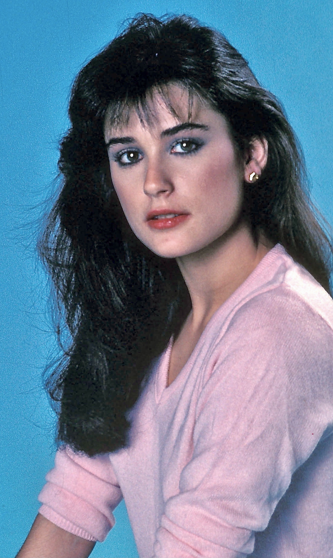 A young Demi Moore gazes softly into the distance, her dark, voluminous hair cascading over a pastel pink sweater that radiates warmth against a cool blue backdrop. Her expression is poised yet tender, framed by subtle makeup that highlights her natural charm.