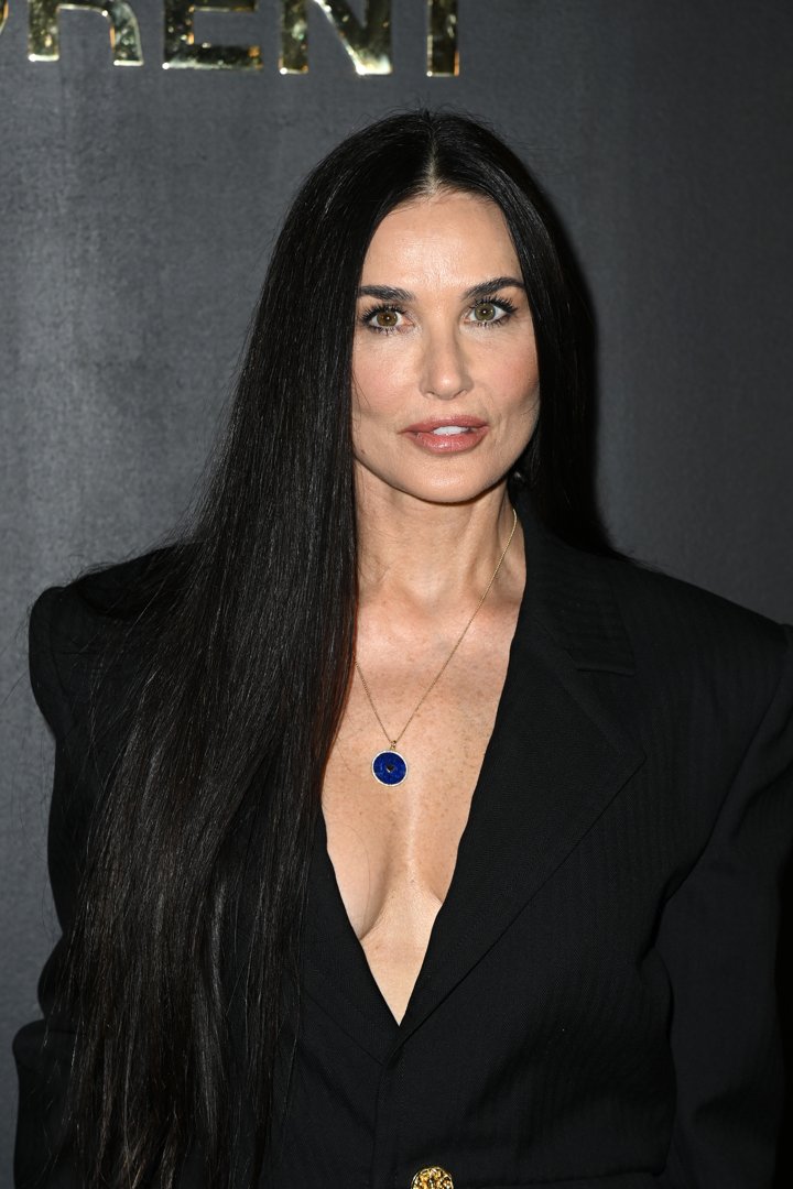 Demi Moore commands attention in a sleek black ensemble, her long, straight hair cascading like silk against the dark backdrop. A deep neckline and a striking blue pendant add a bold touch of contrast and confidence. Her poised expression reflects both strength and timeless sophistication.