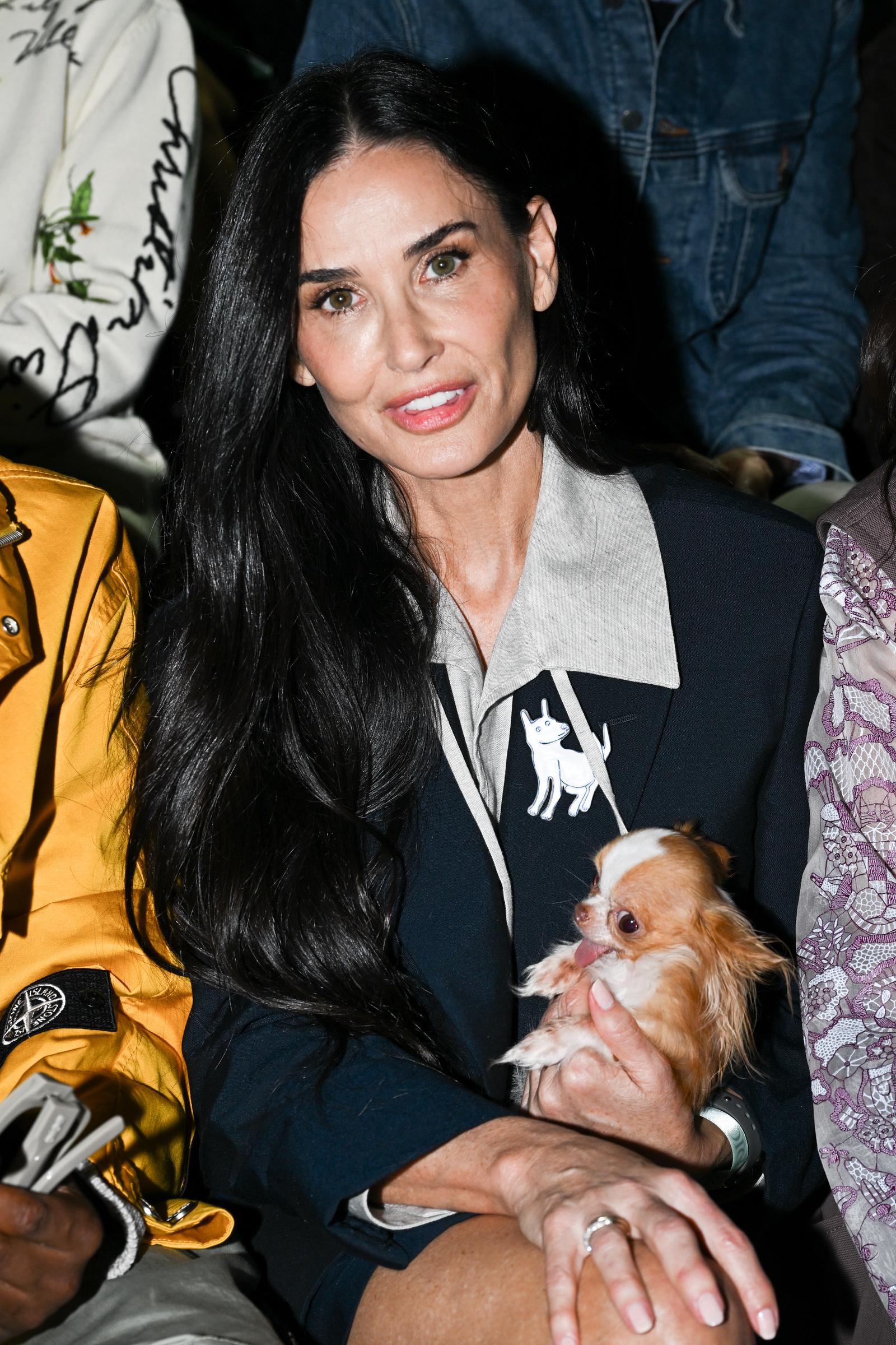 Demi Moore sits poised with a gentle smile, her hair cascading over a chic tailored outfit accented by a playful dog motif. Cradling a tiny dog in her arms, she radiates warmth and approachability. The candid moment captures her blend of elegance, charm, and genuine affection.