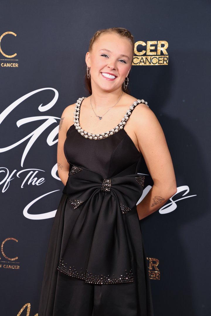 Siwa, also 22, stepped onto the red carpet at the Dancers Against Cancer gala with a completely different kind of glam. Wearing a black satin dress adorned with pearls and a structured bow, she smiled brightly under slick braids and sparkling hoop earrings. Her look was polished, playful, and rooted in performance-ready poise — still high-energy, but with a softer edge.