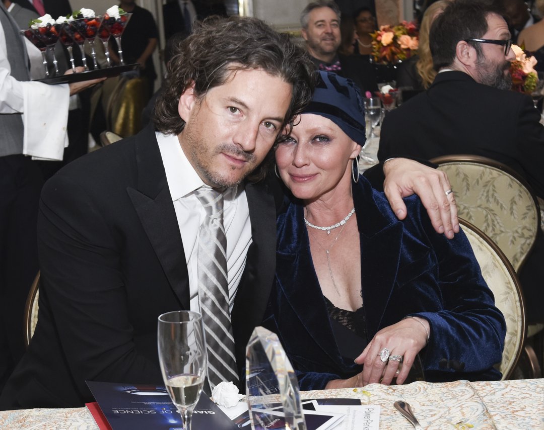 Shannen Doherty and photographer Kurt Iswarienko are pictured seated closely at a formal event, sharing a warm, intimate moment. Dressed in a dark velvet outfit and a headscarf, Doherty leans into Iswarienko, who wears a classic black suit and striped tie in 2016.