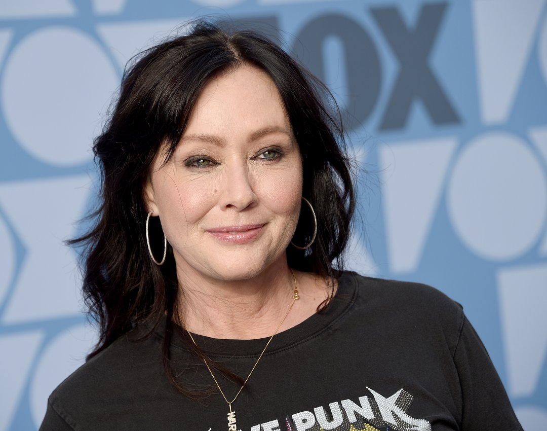 Shannen Doherty is pictured at a FOX event in 2019, wearing a black graphic T-shirt and gold jewelry, with large hoop earrings. Her relaxed smile and softly tousled hair give the portrait a casual, confident tone.