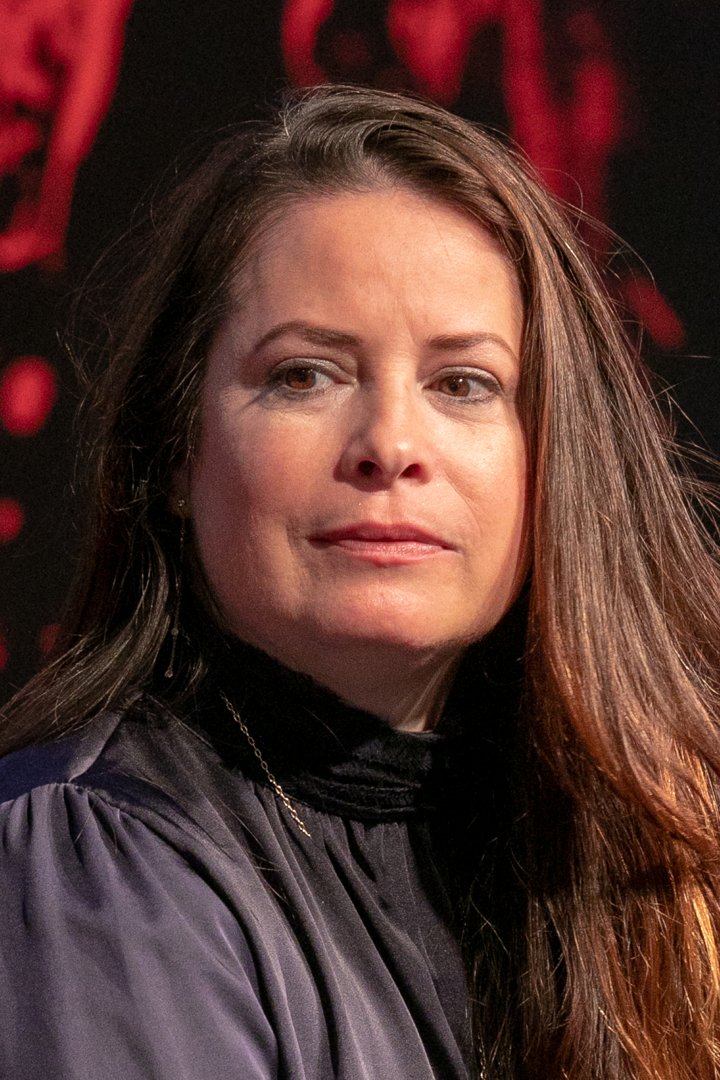 Holly Marie Combs appears at a public event in this close-up taken in 2018. Wearing a high-neck, dark blouse with minimal jewelry, she looks slightly to the side with a composed, thoughtful expression. The red and black backdrop adds contrast to the intimate, candid moment.