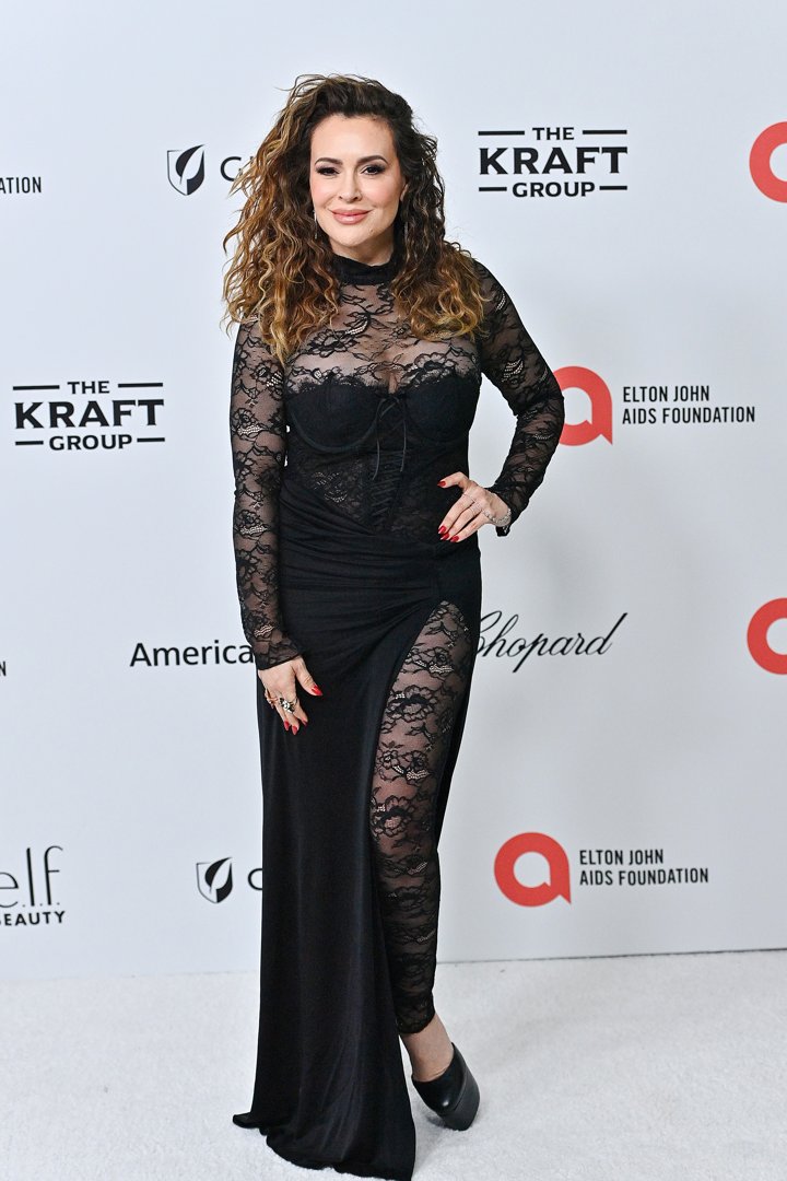Alyssa Milano attends the Elton John AIDS Foundation event in 2024, wearing a striking black gown with sheer lace panels. Her voluminous curls, bold eye makeup, and red nails add to the dramatic elegance of the look. Posing confidently on the white carpet, she brings a sense of glamour to the high-profile occasion.