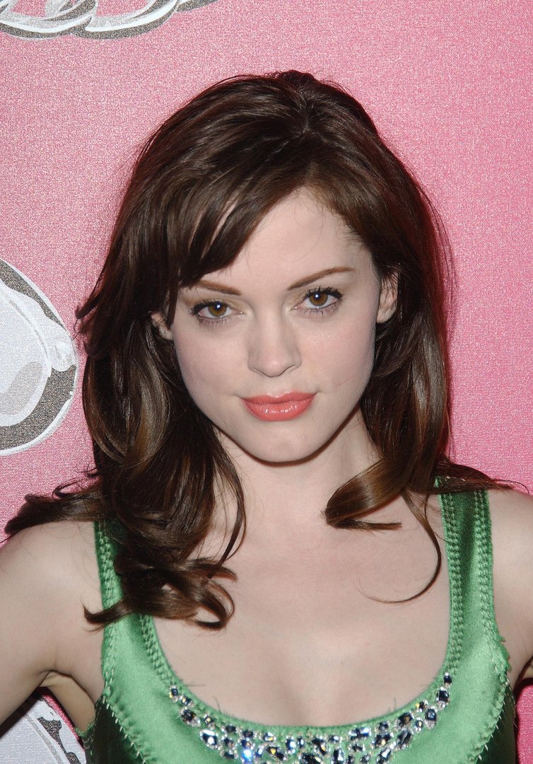 Rose McGowan poses at a red carpet event in the mid-2000s, wearing a satin green dress embellished with crystal detailing at the neckline. Her softly curled hair and pink-toned makeup complement the sparkling pink backdrop, giving the image a vibrant, glamorous feel.