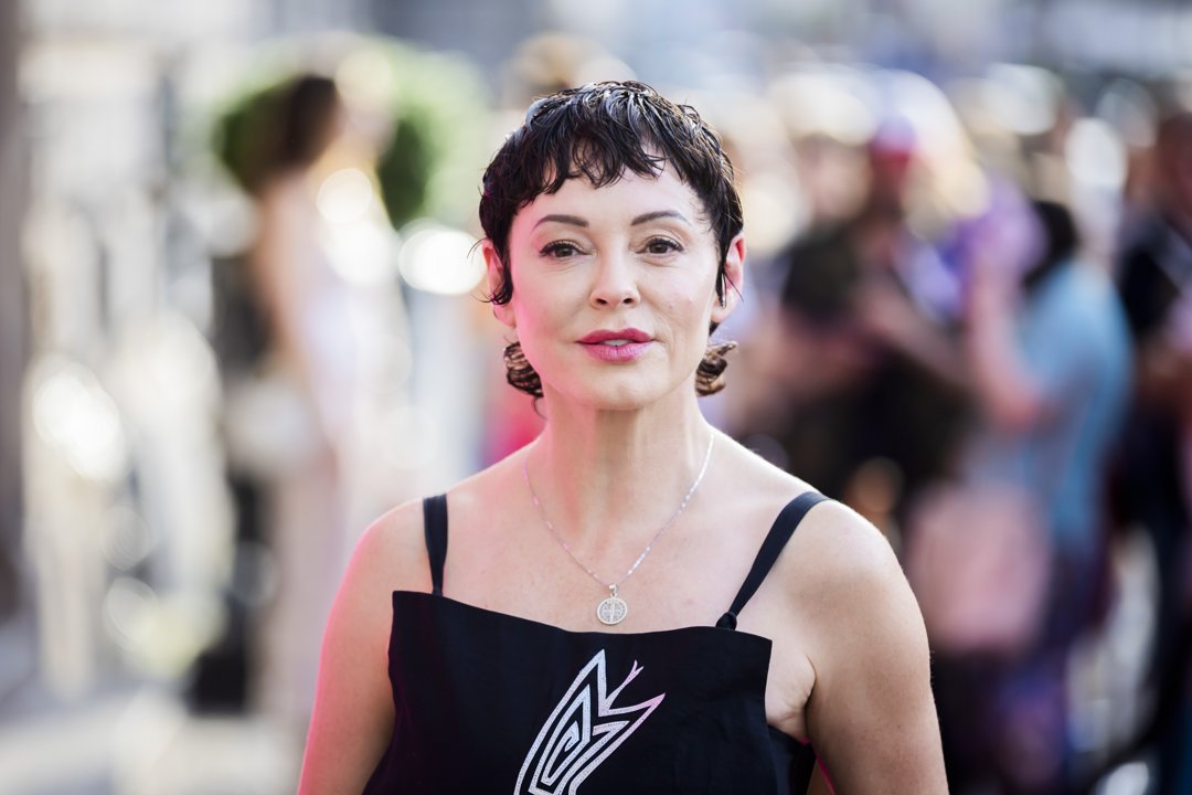 Rose McGowan is seen attending an event in 2024, wearing a black dress with a graphic design and thin straps. Her short, wet-styled hair and bold lipstick give the look a distinctive, avant-garde edge. With a composed expression and a silver pendant necklace, she stands out against the blurred, sunlit crowd in the background.
