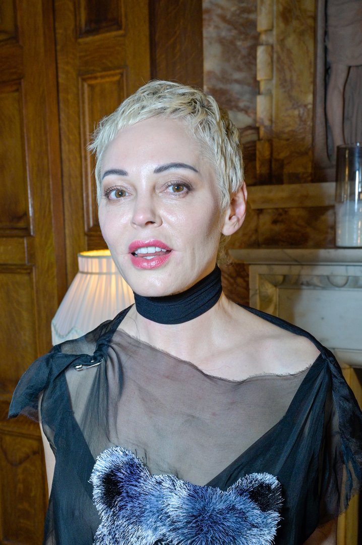 Rose McGowan is photographed indoors in 2025, wearing a sheer black top adorned with a textured bear design and a black choker. Her short platinum-blonde hair and glossy pink lips contrast with the warm wood paneling and soft lighting in the background, creating a dramatic yet intimate portrait.