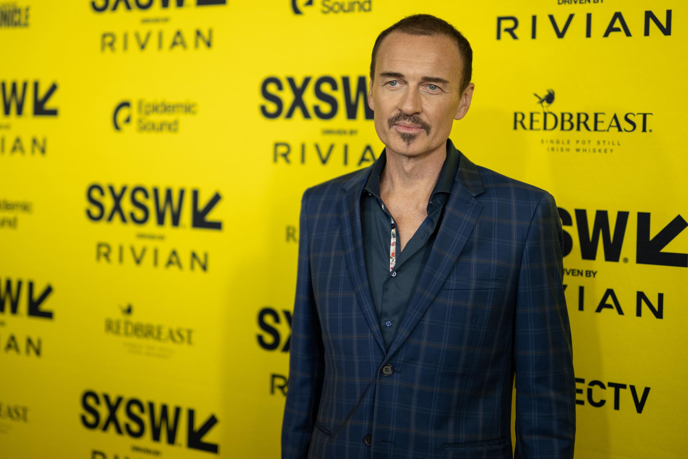 Julian McMahon attends SXSW in 2025, posing in front of a vibrant yellow step-and-repeat backdrop. Wearing a dark blue plaid blazer over a navy shirt with a floral-trimmed collar, he sports a close-cropped hairstyle and goatee. His composed expression adds a sleek, modern edge to the festival appearance.