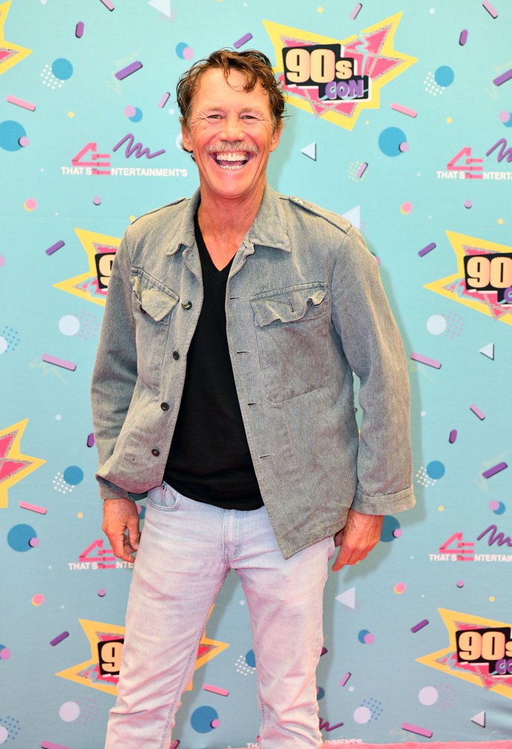 Brian Krause attends 90s Con in 2024, posing on the red carpet with a wide smile. Dressed casually in a light denim jacket, black T-shirt, and pale jeans, he embraces the playful energy of the event. The colorful, retro-themed backdrop enhances the nostalgic tone of the appearance.