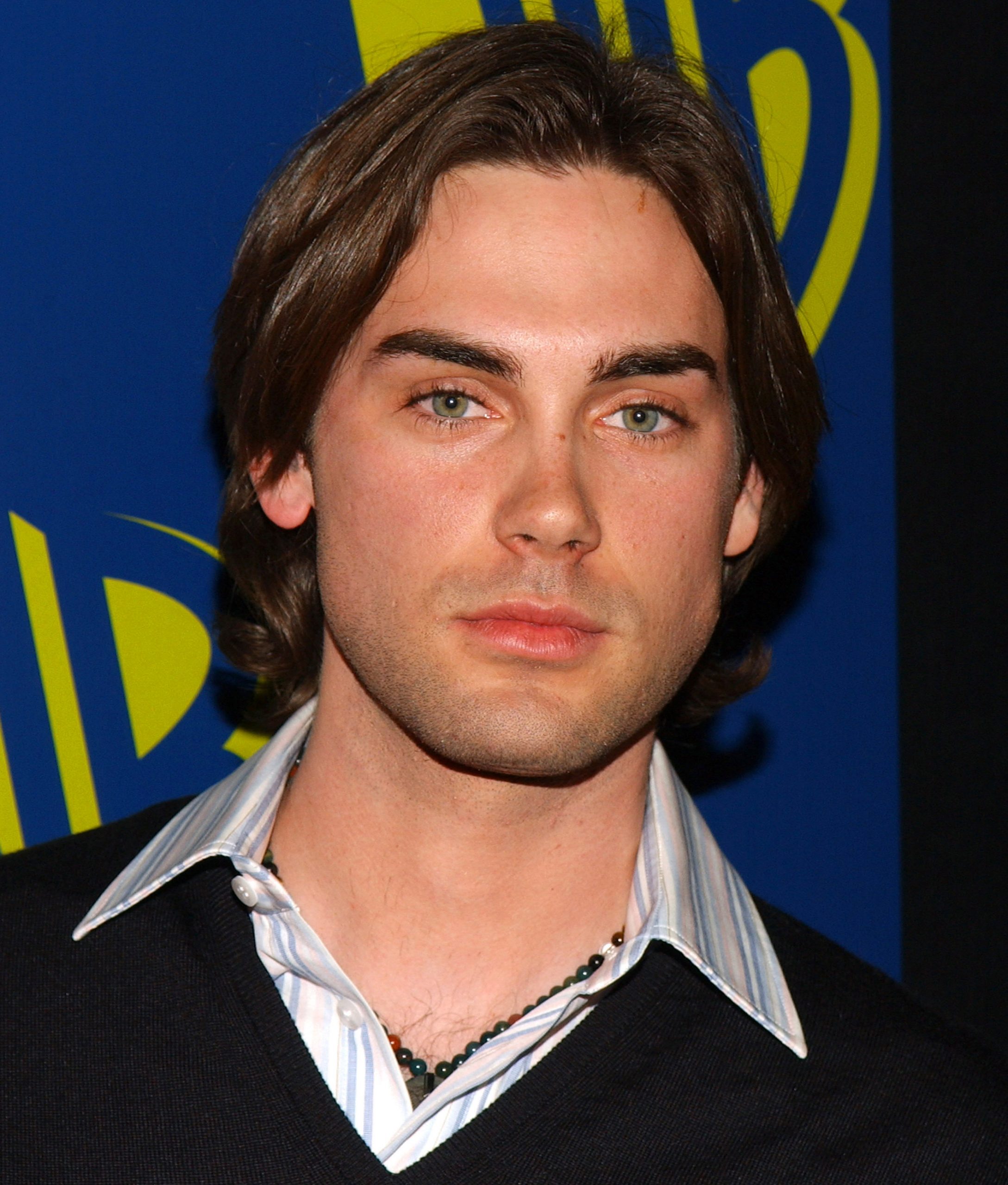 Drew Fuller is seen at a WB network event in the mid-2000s, wearing a striped collared shirt layered under a dark V-neck sweater. His shoulder-length hair and serious expression give the portrait a polished, youthful intensity, fitting for his role during his time on