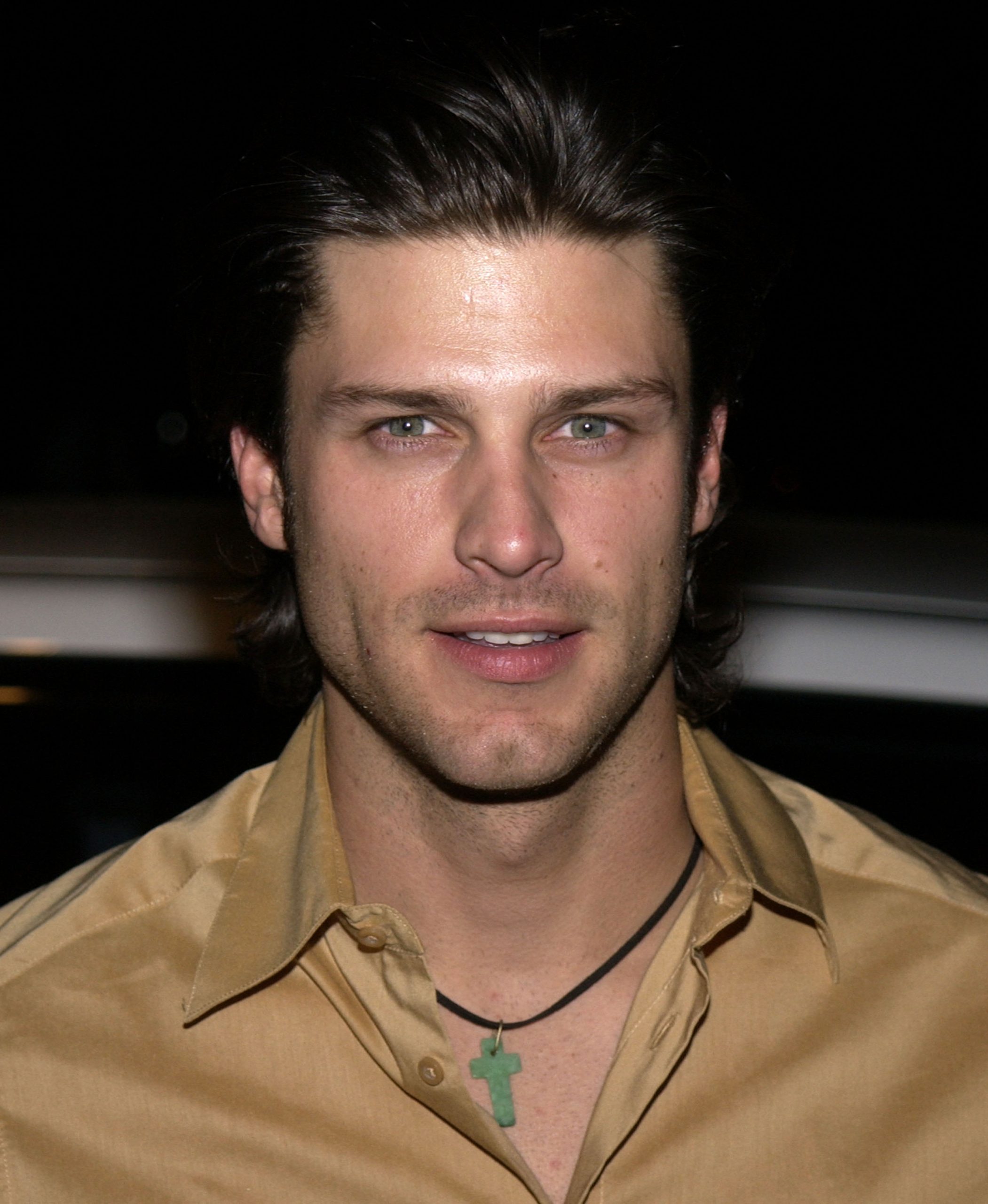 Greg Vaughan is photographed at a red carpet event in the early 2000s, wearing a gold button-up shirt and a green cross pendant necklace. His hair is swept back in a classic style, and his direct gaze, combined with the soft flash lighting, gives the image a polished yet casual feel.