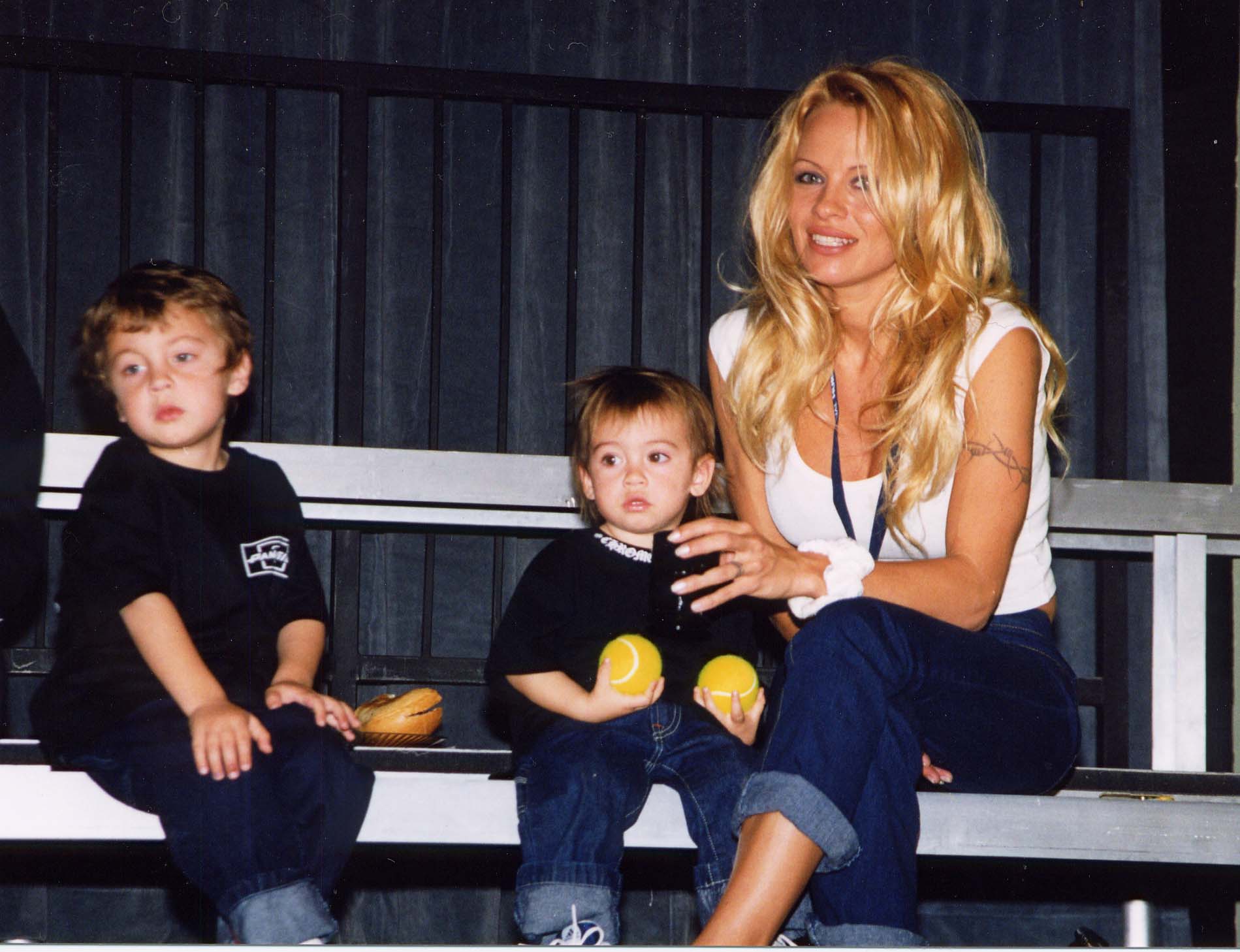 Anderson sits beside her young sons, Brandon and Dylan, during a casual public outing in 1999. Dressed in a white tank top and rolled-up jeans, Anderson smiles warmly while holding Dylan, who clutches two yellow tennis balls. Brandon, seated just beside them in a matching black shirt and jeans, gazes off to the side. The intimate moment, captured on metal bleachers, offers a glimpse into the early years of the Anderson-Lee family.