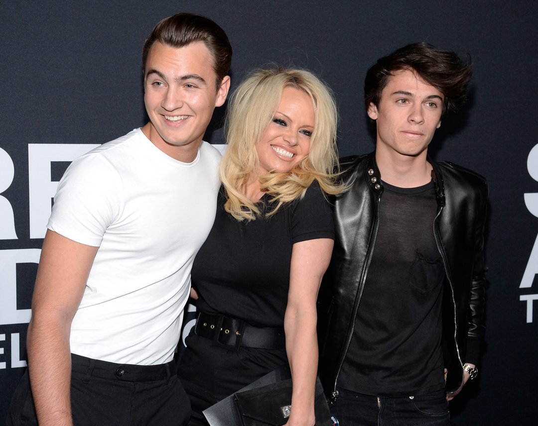 Anderson beams as she poses with Brandon and Dylan, at a 2016 red carpet event. Wearing a fitted black dress cinched with a wide belt, Anderson stands between her sons — Brandon in a crisp white T-shirt and black trousers, and Dylan in a black leather jacket layered over a sheer shirt. The trio's coordinated black-and-white looks highlight their close bond and striking family resemblance.