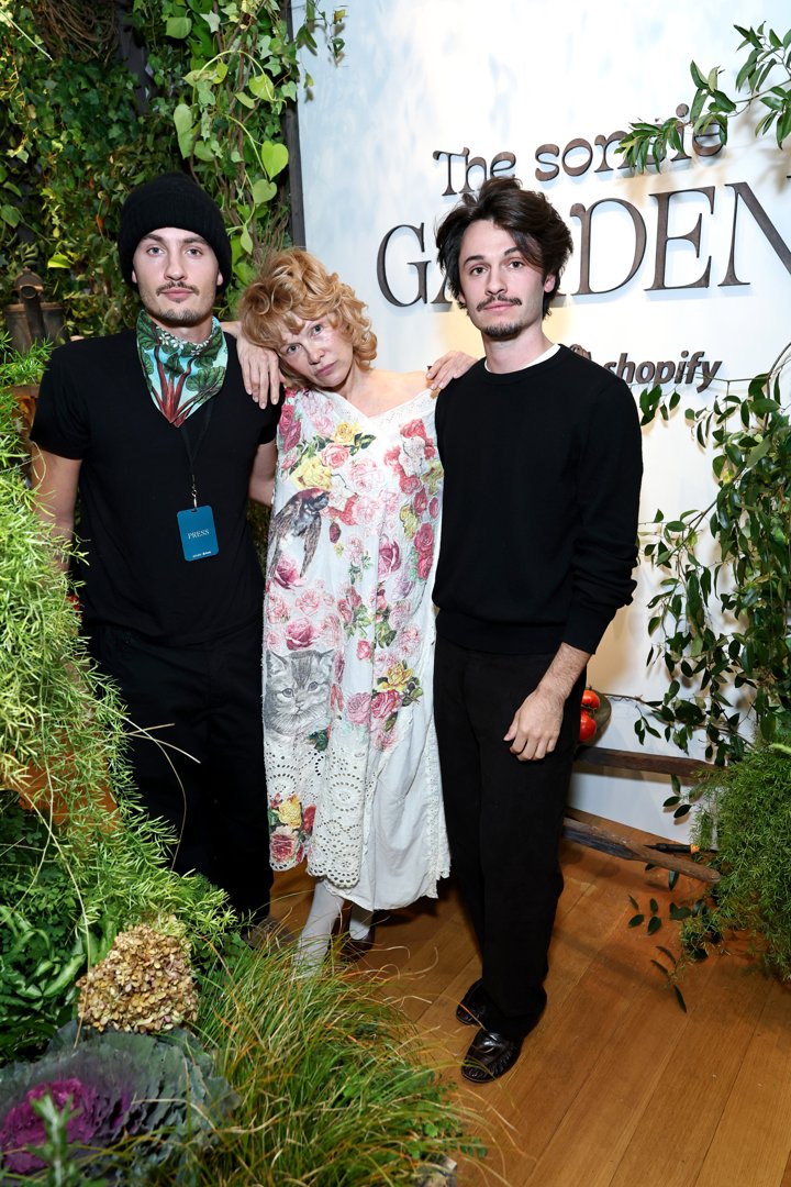 The trio at the Sonsie Garden event in New York City in 2025. Wearing a white floral dress detailed with birds, cats, and roses, Anderson stands with her arms gently draped around her sons. Dylan wears a black beanie and printed scarf, while Brandon opts for a simple black crewneck. Surrounded by greenery and natural textures, the trio presents a striking image of family presence and quiet unity.