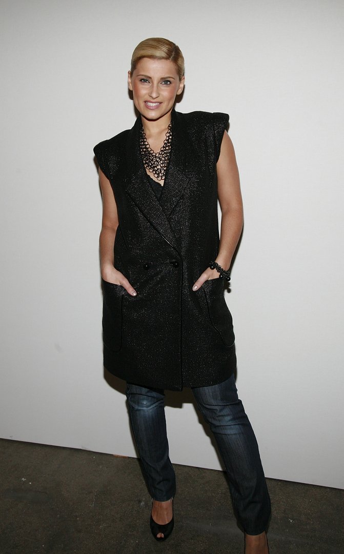 Furtado poses in a sleeveless black textured blazer with a double-breasted design, paired with dark blue jeans and black peep-toe heels. Her hair is styled in a sleek, side-parted updo, and she accessorizes with a layered black beaded necklace and matching bracelet.