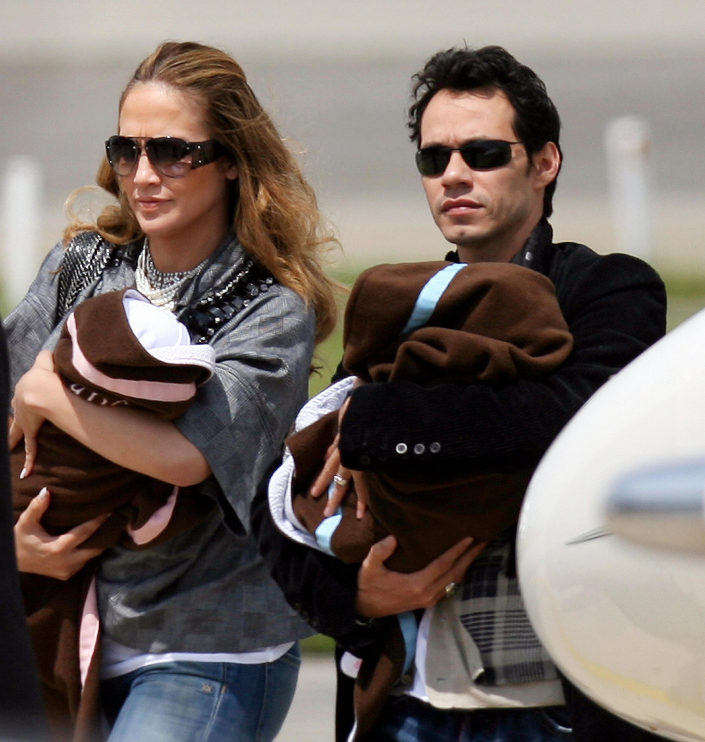 Jennifer Lopez and Marc Anthony, then still married, arrive in Zaventem with their twins. Each parent cradled one of the babies, wrapped in matching blankets — Jennifer holding a pink one, likely Emme, and Marc carrying a blue one, presumably Max.
