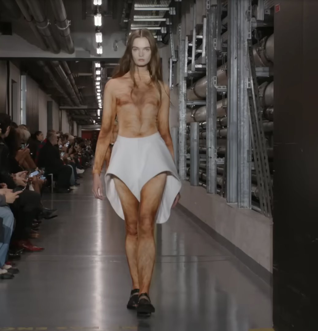 Another male model made a bold entrance in a striking white skirt with asymmetric high-and-low sections. It appears to be his only piece of clothing as he confidently strides down the runway. From a post dated October 5, 2025. Source: Youtube/@jeanpaulgaultier