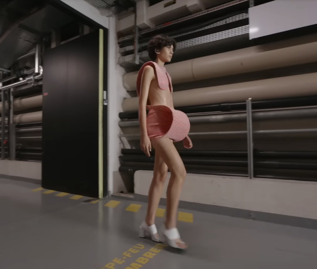 Another standout design is worn by this model, styled with white block heels. The top appears to rest lightly on his shoulders, leaving part of the upper body exposed, while the skirt features a uniquely structured, sculptural build at the front. From a post dated October 5, 2025. Source: Youtube/@jeanpaulgaultier