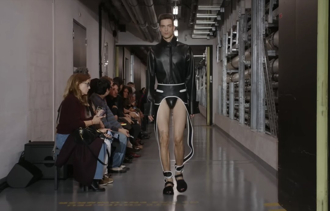 A model walks by in a striking black, groin-baring outfit. The design resembles a coat but features side detailing that extends down the legs, along with a fitted bodysuit section at the center. From a post dated October 5, 2025. Source: Youtube/@jeanpaulgaultier