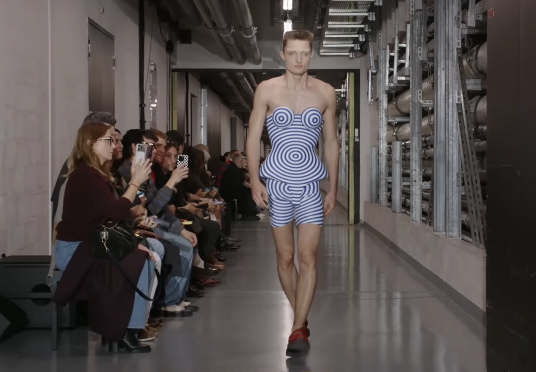 A model showcases another futuristic design that appears to feature a 3D-printed effect. The peplum-shaped top is paired with matching shorts, creating a daring and modern look on the male model. From a post dated October 5, 2025. Source: Youtube/@jeanpaulgaultier