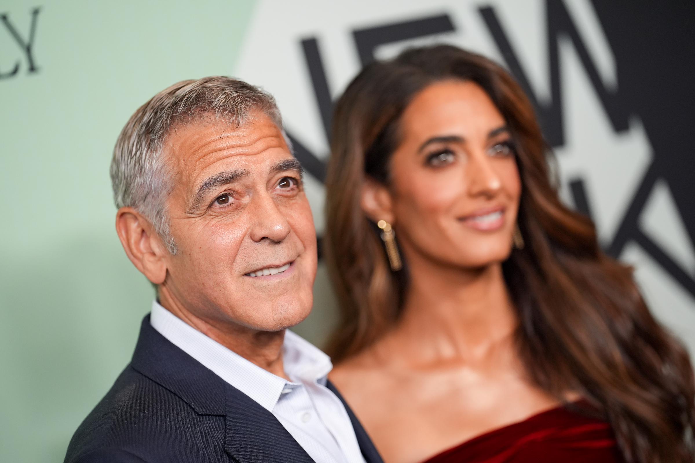 On the red carpet, George Clooney watches on as Amal Clooney poses with ease. The floral texture of her dress offers a bold contrast to his clean, classic look.
