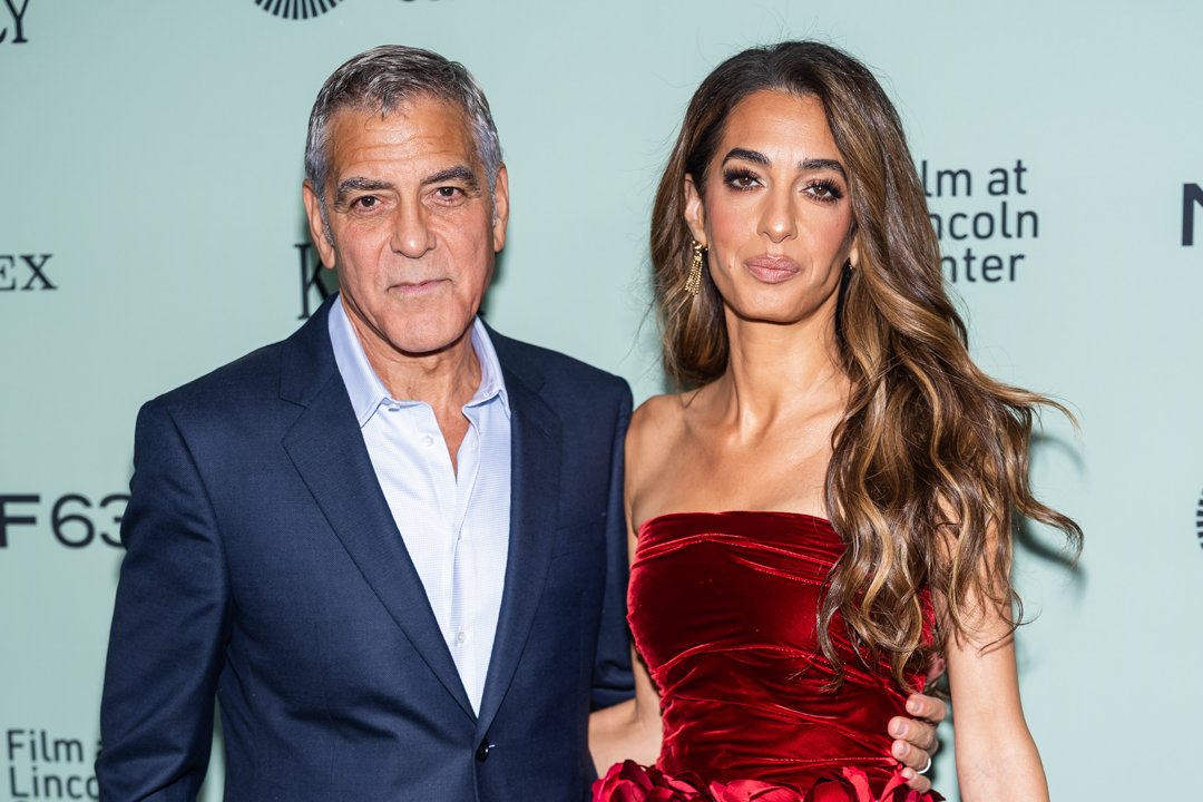 George Clooney and Amal Clooney face the press wall side by side. She offers a faint, composed smile while he watches her with a quiet, unmistakably proud expression.