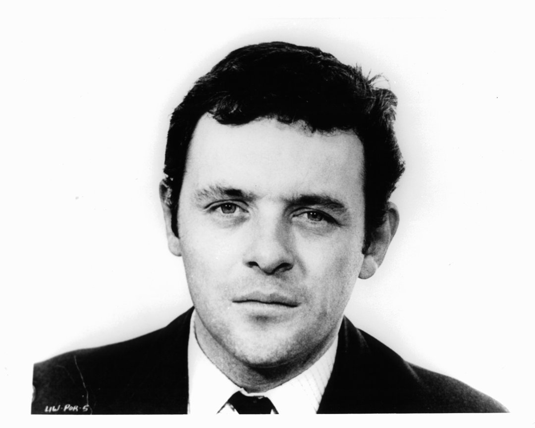 A Black-and-white photograph that captures a young Anthony Hopkins with an intense yet contemplative expression. His gaze is steady and direct, conveying both confidence and quiet introspection. The lighting accentuates the texture of his hair and the subtle shadowing around his face, creating a timeless, cinematic quality.