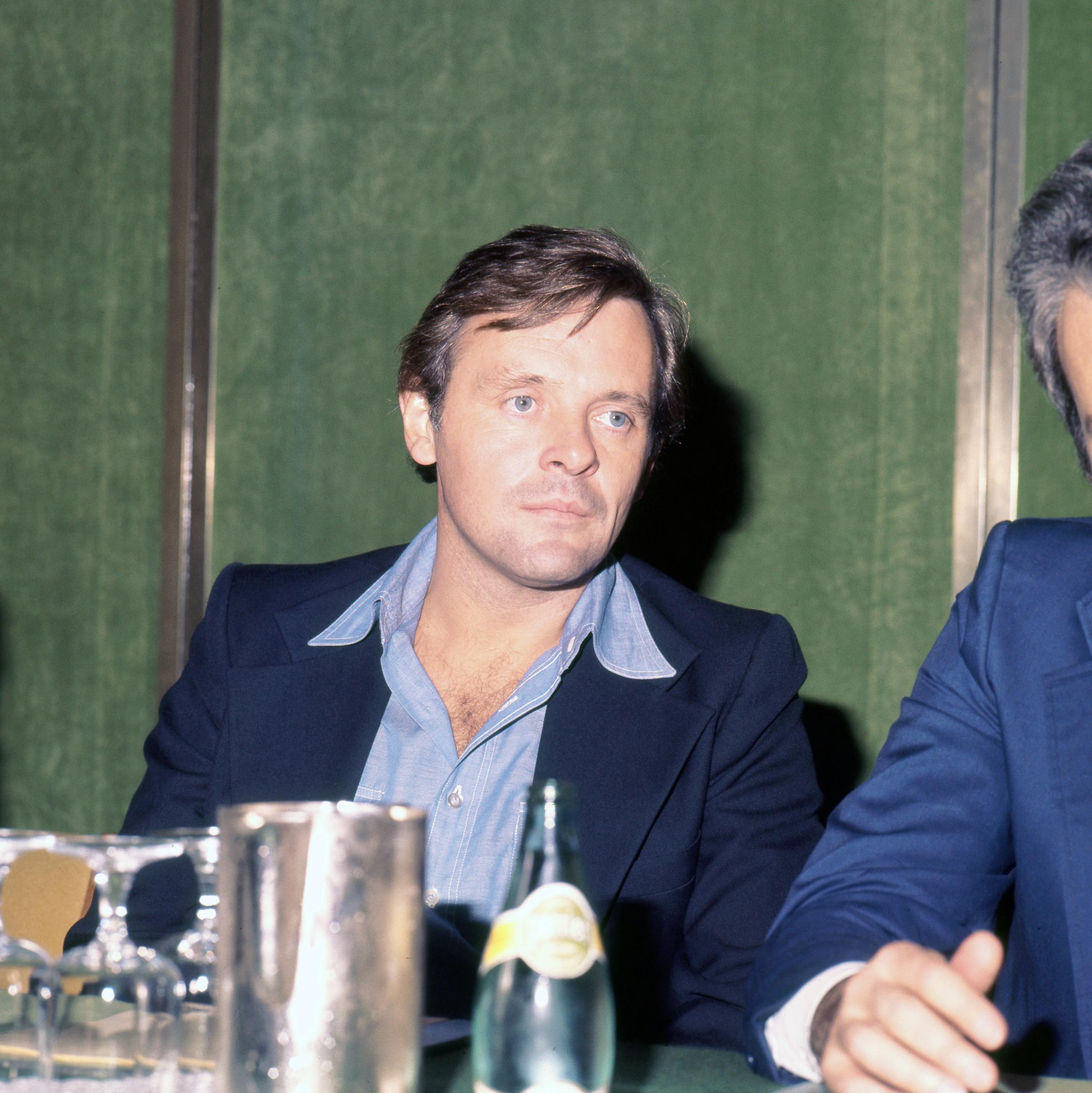 Sitting poised at a press conference table in London, Anthony Hopkins's calm demeanor hints at quiet introspection. Dressed in a crisp blue shirt and tailored jacket, he radiates effortless sophistication. The soft lighting and reflective glassware lend the scene a timeless air of composure and grace.