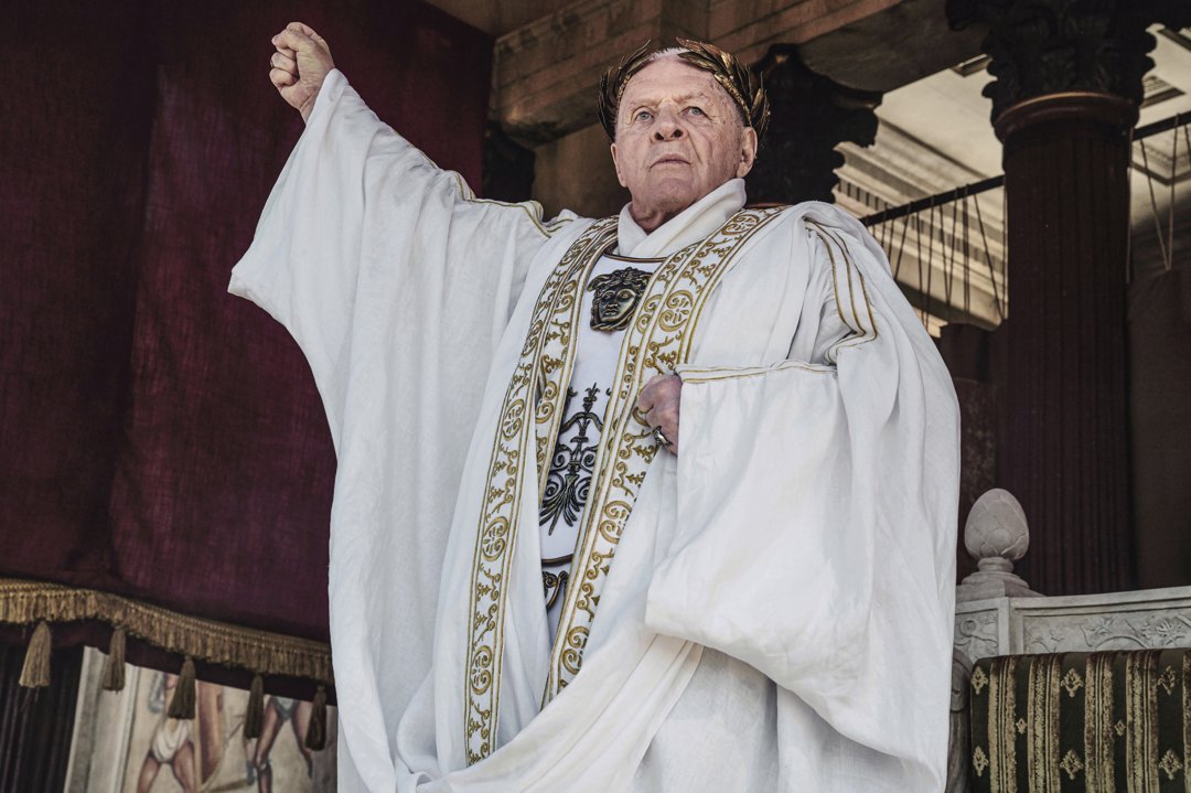 Commanding the scene as Emperor Vespasian while draped in regal white robes adorned with golden embroidery, Anthony Hopkins captures the grandeur and gravitas of an emperor immortalized in history. His raised arm and resolute gaze embody the power and authority of Rome's might.