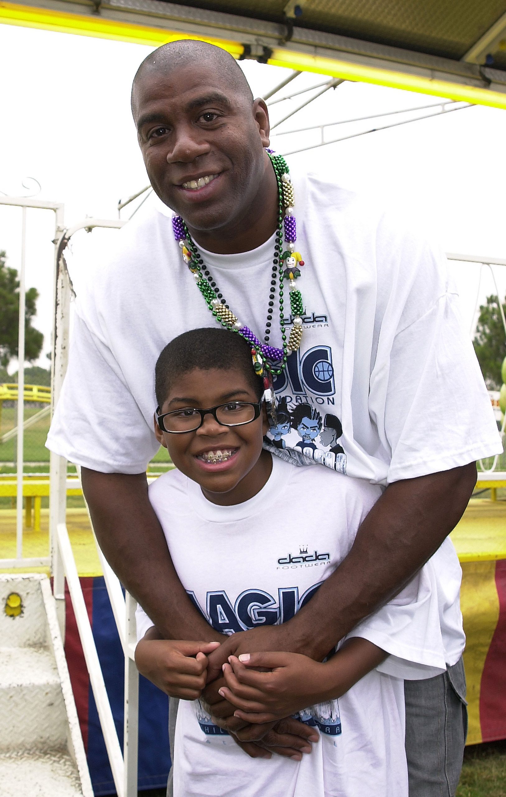 Beaming with pride, Magic Johnson wraps his arms around a young EJ Johnson during the Magic Johnson Foundation's Mardi Gras event, radiating joy at a day built for family and community.