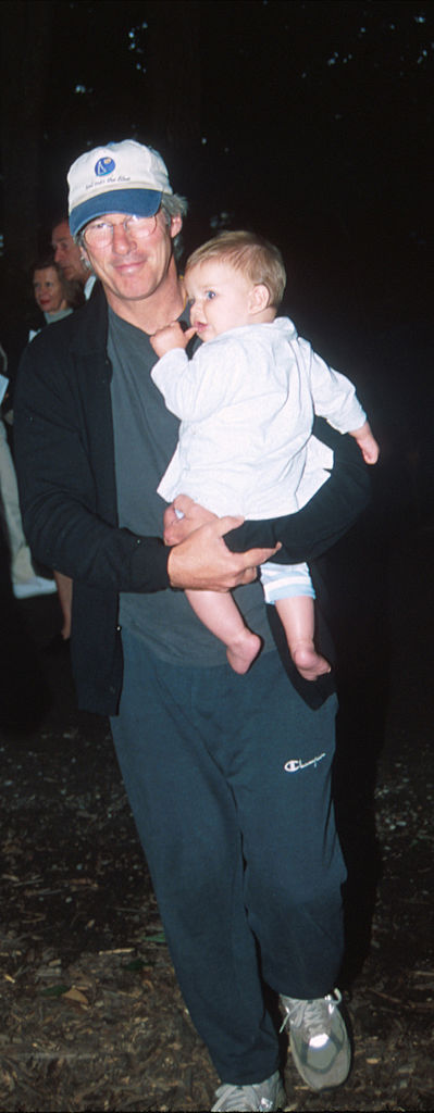 Richard Gere flashes a warm smile as he carries baby Homer Gere in his arms at the Watermill Center’s Summer Gala Benefit in Water Mill, New York — a quiet, affectionate father-son moment in the spotlight.