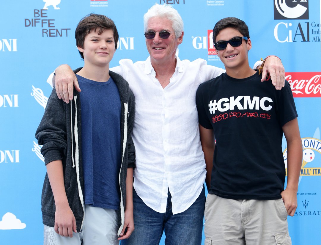 Homer Gere stands casually with a soft smile beside his father Richard Gere and a friend at the Giffoni Film Festival in Italy. With arms draped over shoulders, the trio strikes a laid-back pose in front of the event backdrop.