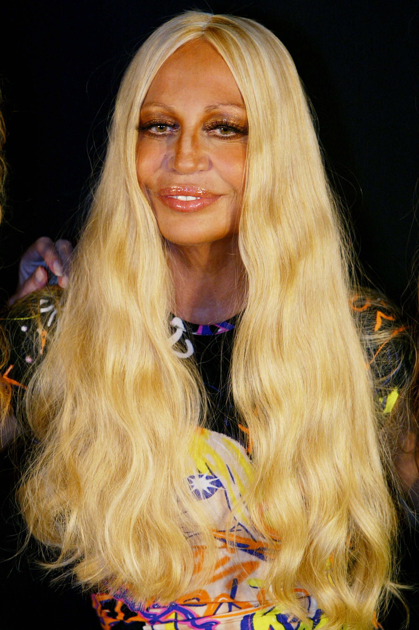 Donatella Versace, 48, smiled before Gianni Versace's show on October 4, 2003, in Milan, Italy. Her face displayed extreme tightness, heavily augmented, glossy lips, and a deeply tanned skin tone.