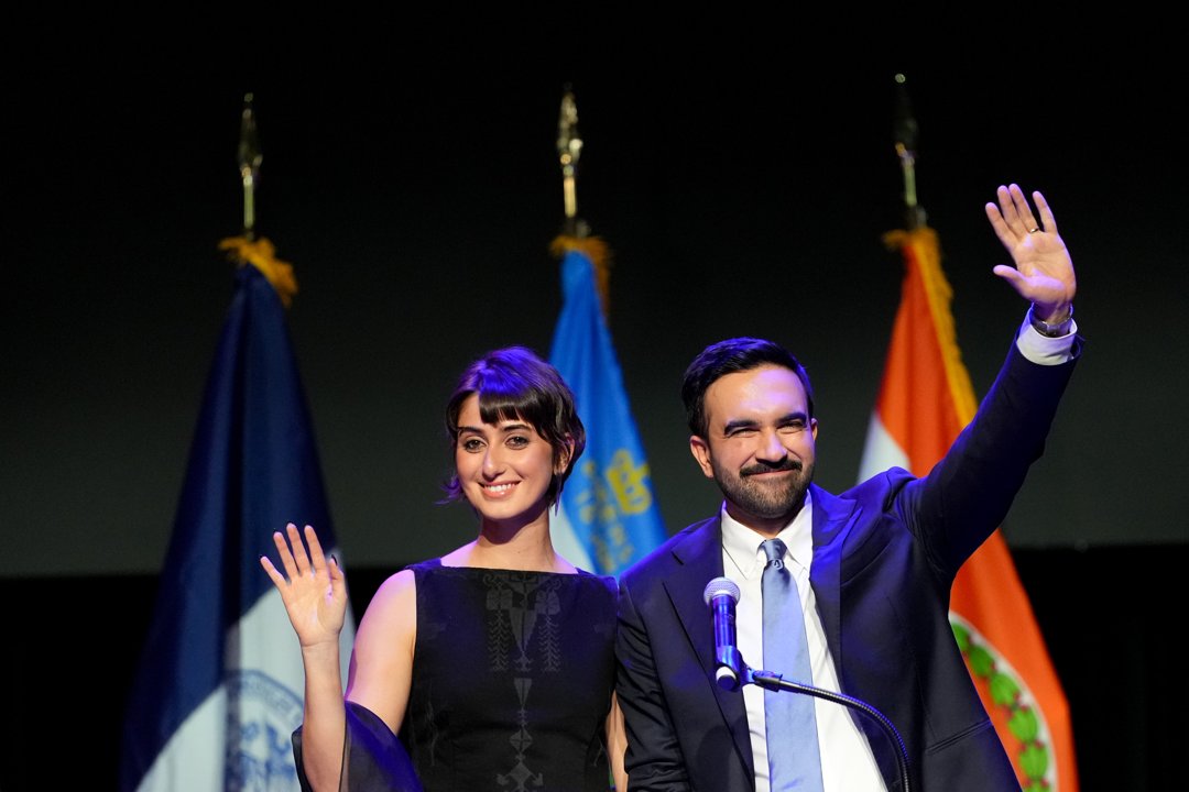 Beaming with pride, Zohra Mamdani and Rama Duwaji wave to the crowd from the stage, framed by city and state flags during a night of celebration and victory in Brooklyn.