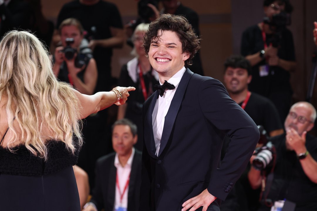 Wearing a tux and glowing with confidence, Homer Gere takes center stage on the red carpet at the 81st Venice International Film Festival. He smiles brightly as cameras flash and a woman gestures excitedly in his direction.