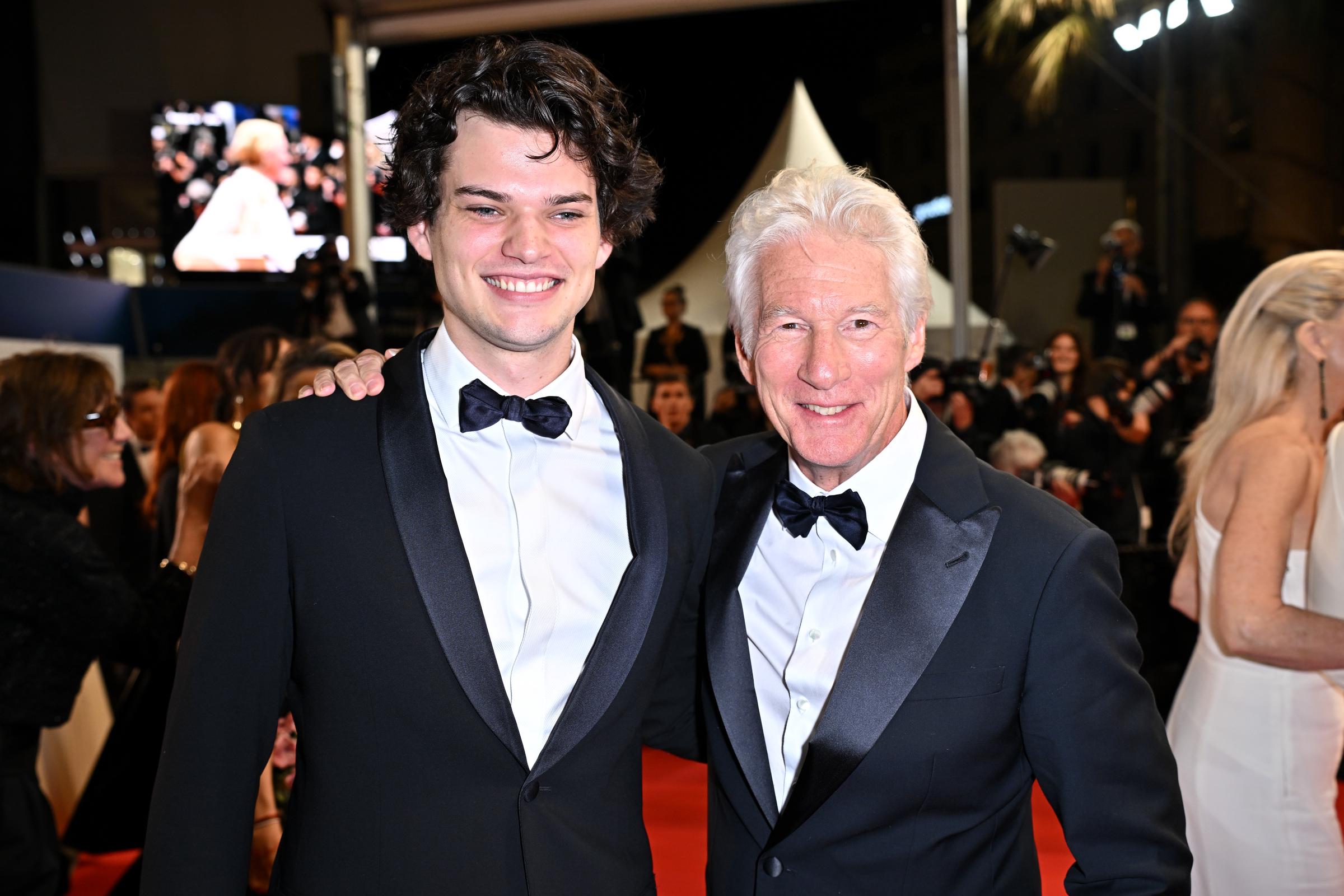 Homer and Richard Gere share a proud red carpet moment at the 77th Cannes Film Festival. Dressed in matching tuxedos, they smile side by side amid a swirl of flashing lights and elegant crowds at the 