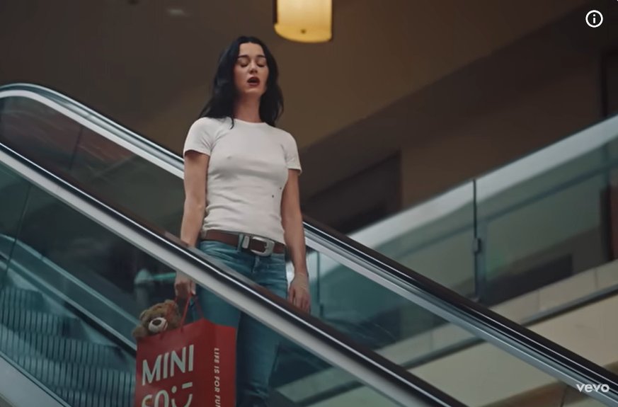Katy Perry stands on a moving escalator, while carrying a shopping bag with a teddy bear inside. | Source: Youtube/KatyPerry