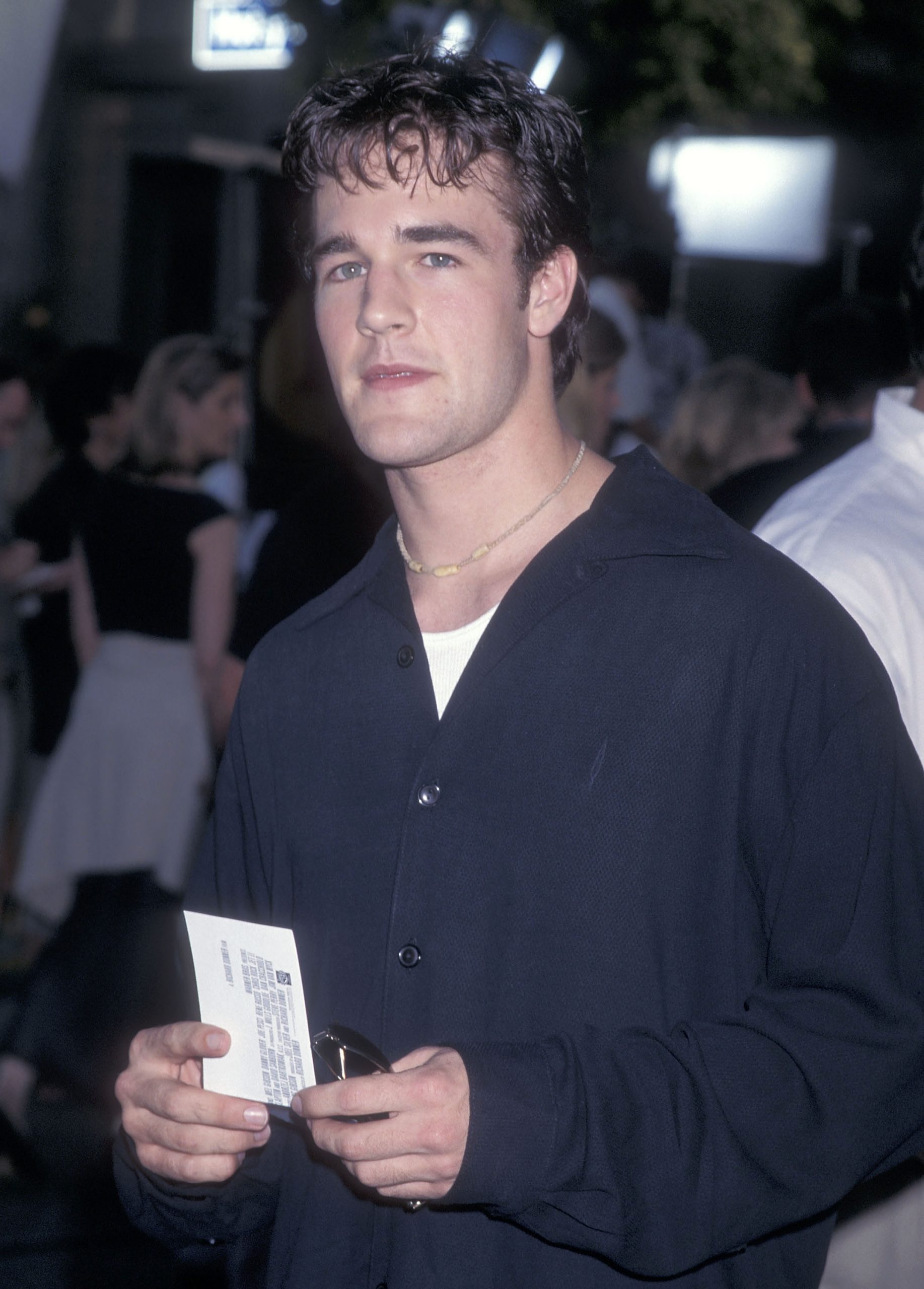 James Van Der Beek stands amid the premiere crowd, holding a ticket as soft lights illuminate the scene around him. His dark shirt and simple necklace give him a relaxed, understated style against the lively backdrop.