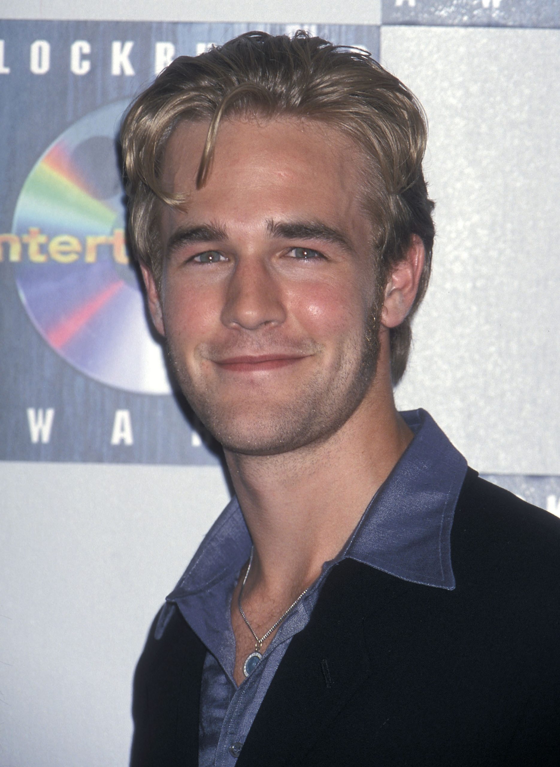 James Van Der Beek arrives at the Sixth Annual Blockbuster Entertainment Awards with a relaxed grin and tousled blond hair, radiating charm. He wears a slate-blue shirt with a wide collar under a dark jacket, softened by a simple silver necklace, exuding polished, youthful confidence.