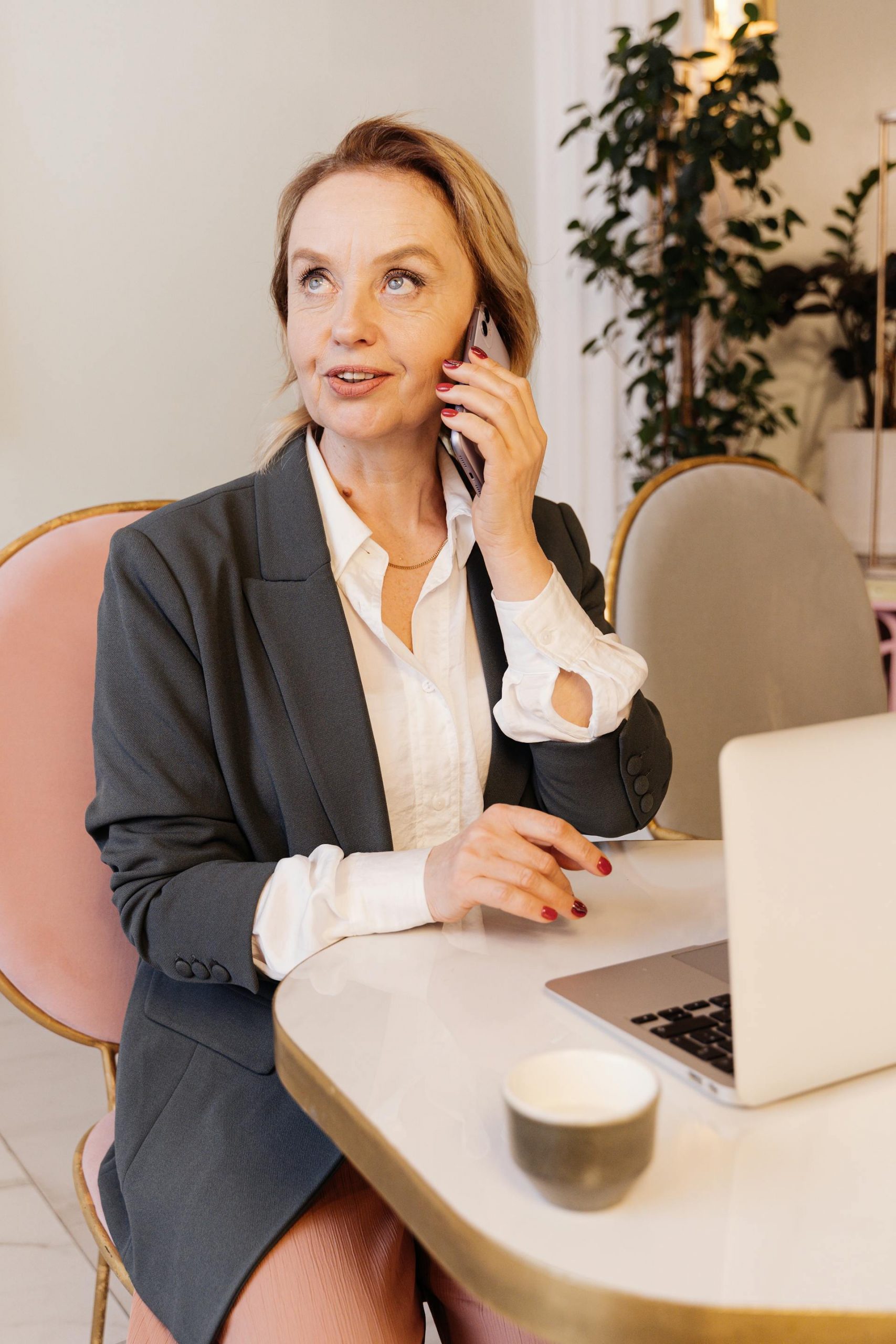 A mature businesswoman in a gray blazer talking on a phone while working on a laptop at a café | Source: Pexels