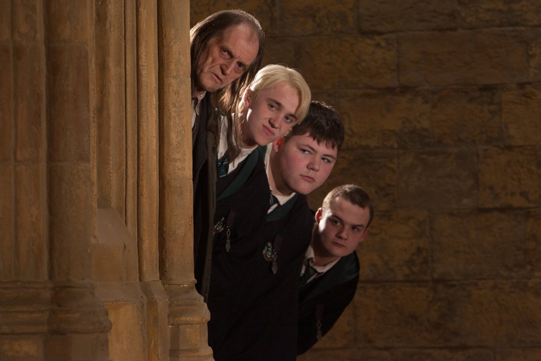 David Bradley, Tom Felton, Jamie Waylett, and Josh Herdman peer around a stone pillar like a mischievous Slytherin totem pole in motion. Their expressions range from sly to suspicious, each one perfectly in character against the cold Hogwarts walls. It's a moment of on-set camaraderie wrapped in classic villainous curiosity from 