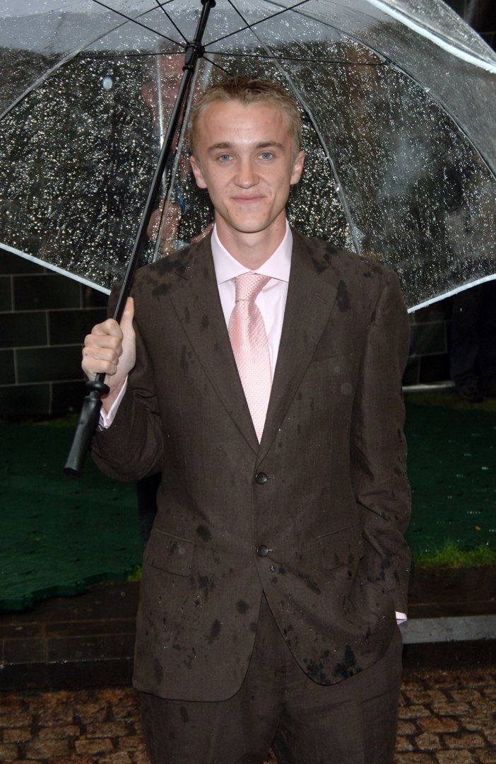 Tom Felton stands under a clear umbrella, rain-speckled and smiling as if the storm were part of the event's charm. His suit bears the marks of London's drizzle, yet he carries himself with easy confidence and good humor. It's a quintessentially British premiere moment and the magic goes on.