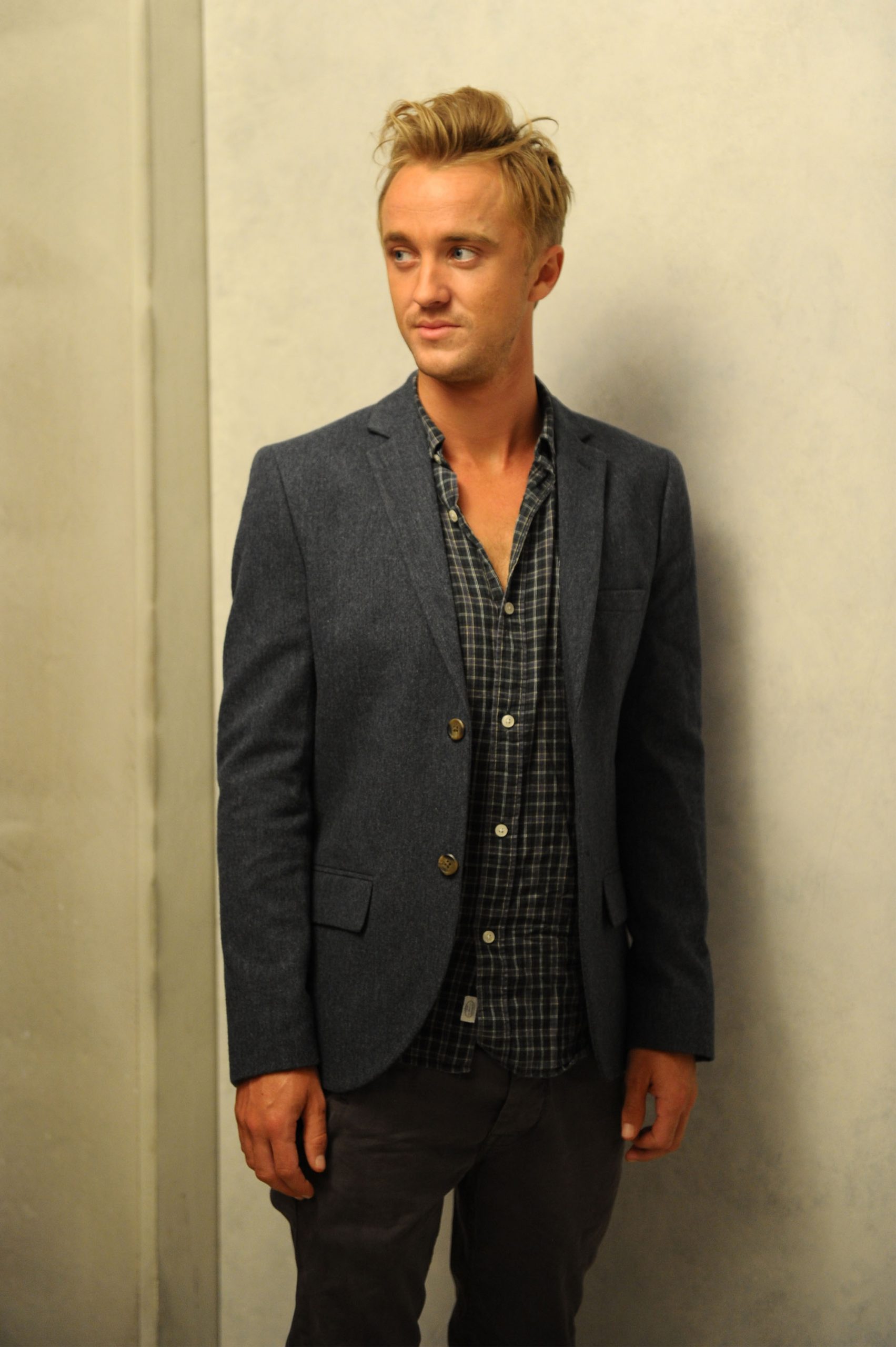 Tom Felton stands in a softly lit studio, blending casual ease with tailored polish in a checked shirt and blue blazer. His relaxed pose and thoughtful glance lend a quiet charm to the Toronto International Film Festival's bustling atmosphere. Amid the event's cinematic excitement, he brings calm, understated confidence.