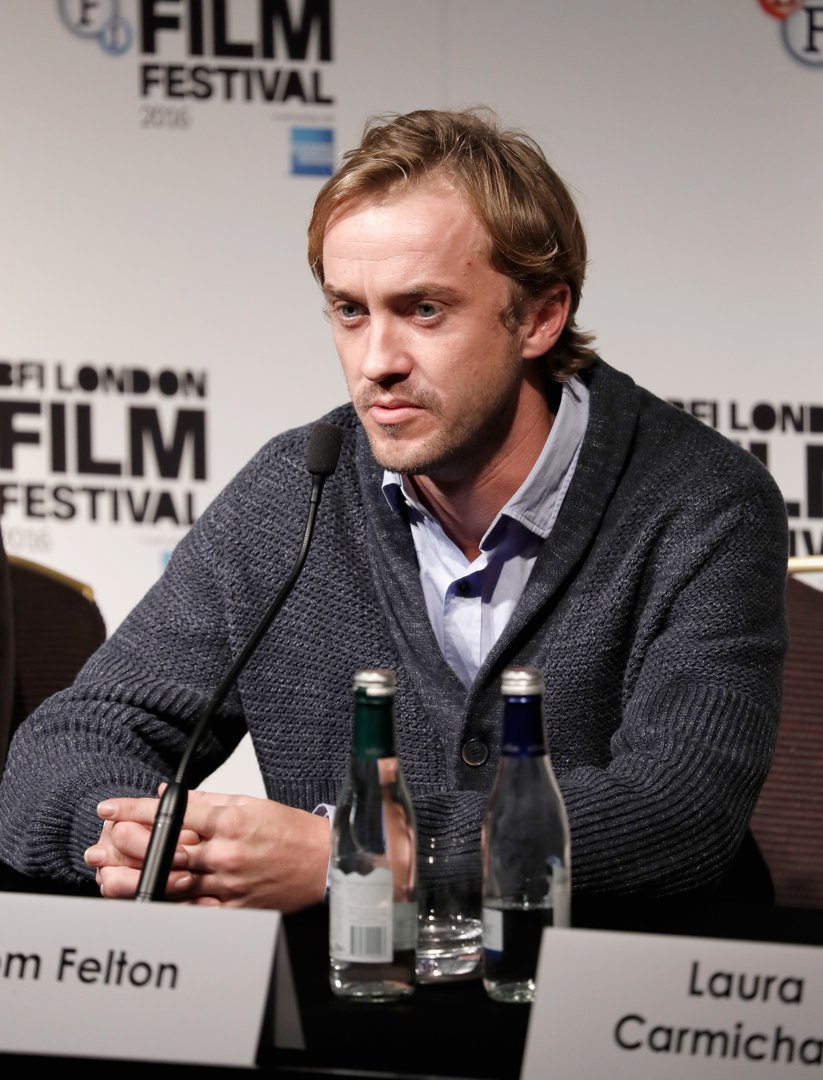 Tom Felton sits poised at the press table, his thoughtful expression matching the weight of the conversation at hand. Wrapped in a textured sweater, he brings a calm, grounded presence to the room of eager listeners. At the 