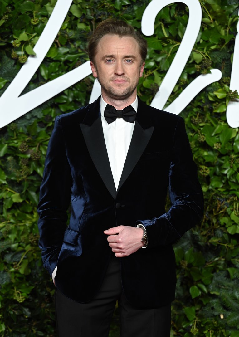 Tom Felton cuts a refined figure in a midnight-blue velvet tuxedo, standing poised against a lush backdrop of greenery. With a classic bow tie and composed expression, he brings understated elegance to the Royal Albert Hall.