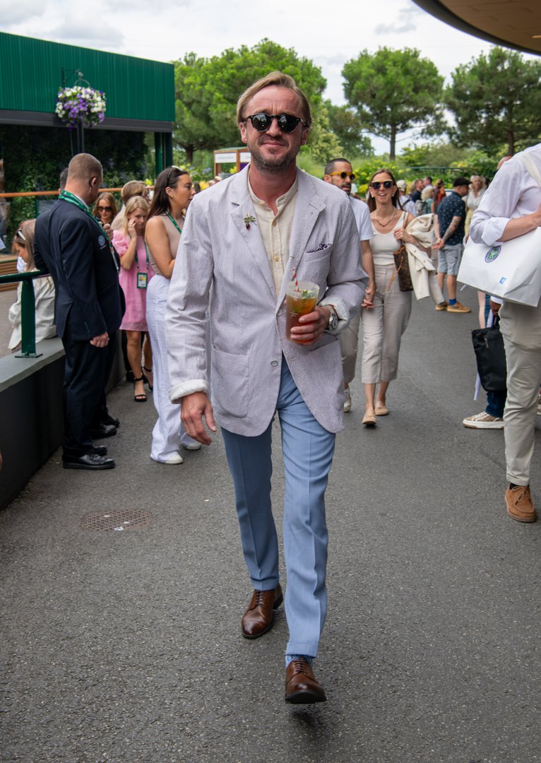 Tom Felton strolls through the Wimbledon crowds with a chilled drink in hand, blending relaxed elegance with summer charm. His light blazer, pastel trousers, and easy confidence capture the spirit of the iconic 